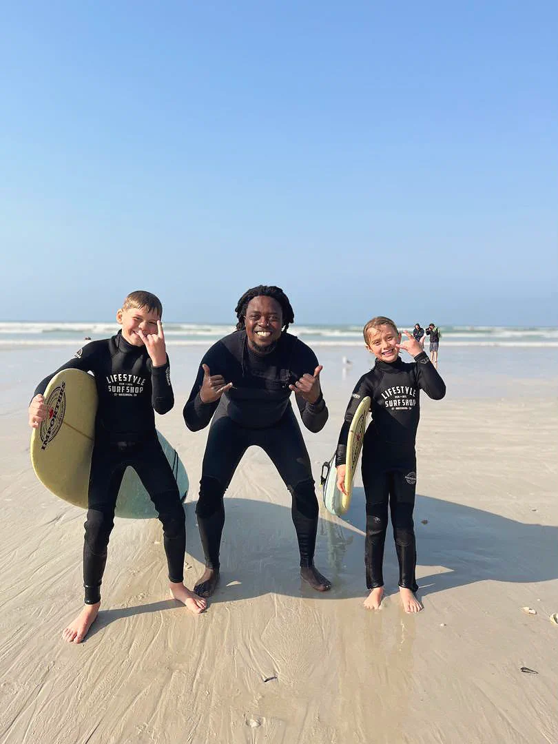 Kwezi on the sand at Muizenberg with two young students in wetsuits, all throwing shakas