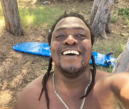 Kwezi, smiling, with his surfboard on the grass behind him